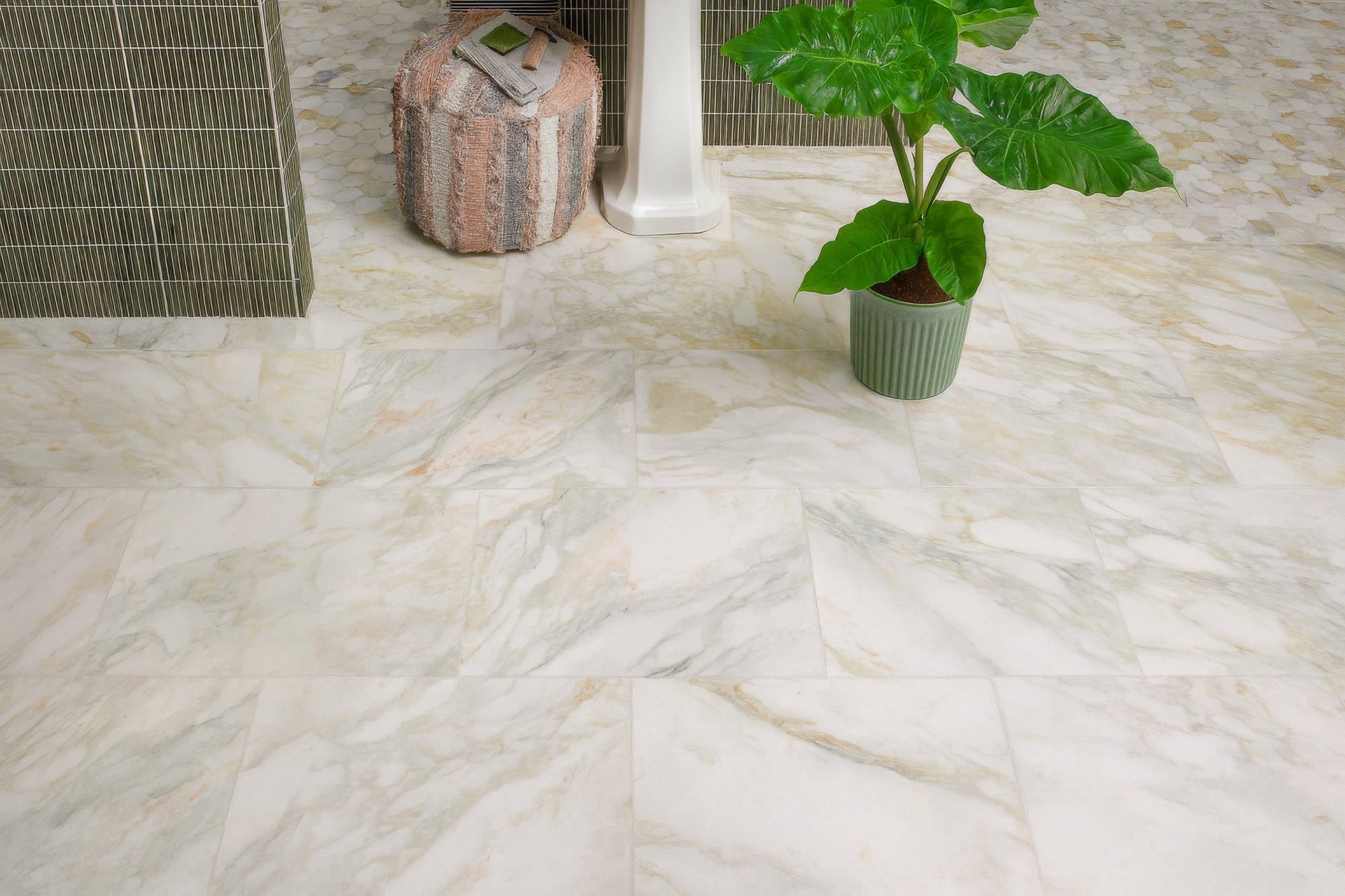 A potted plant with broad green leaves in a textured sage green ceramic pot displayed with marble bathroom tiles