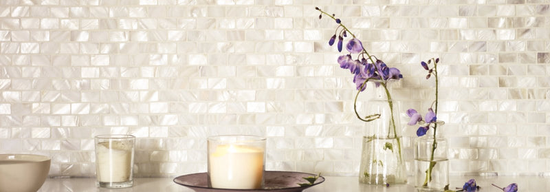 A delicate arrangement of purple flowers in clear glass vases on a countertop with a shimmering white mosaic tile backdrop