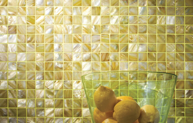 A translucent green glass bowl filled with lemons in front of a shimmering yellow and white mother of pearl mosaic tile backsplash