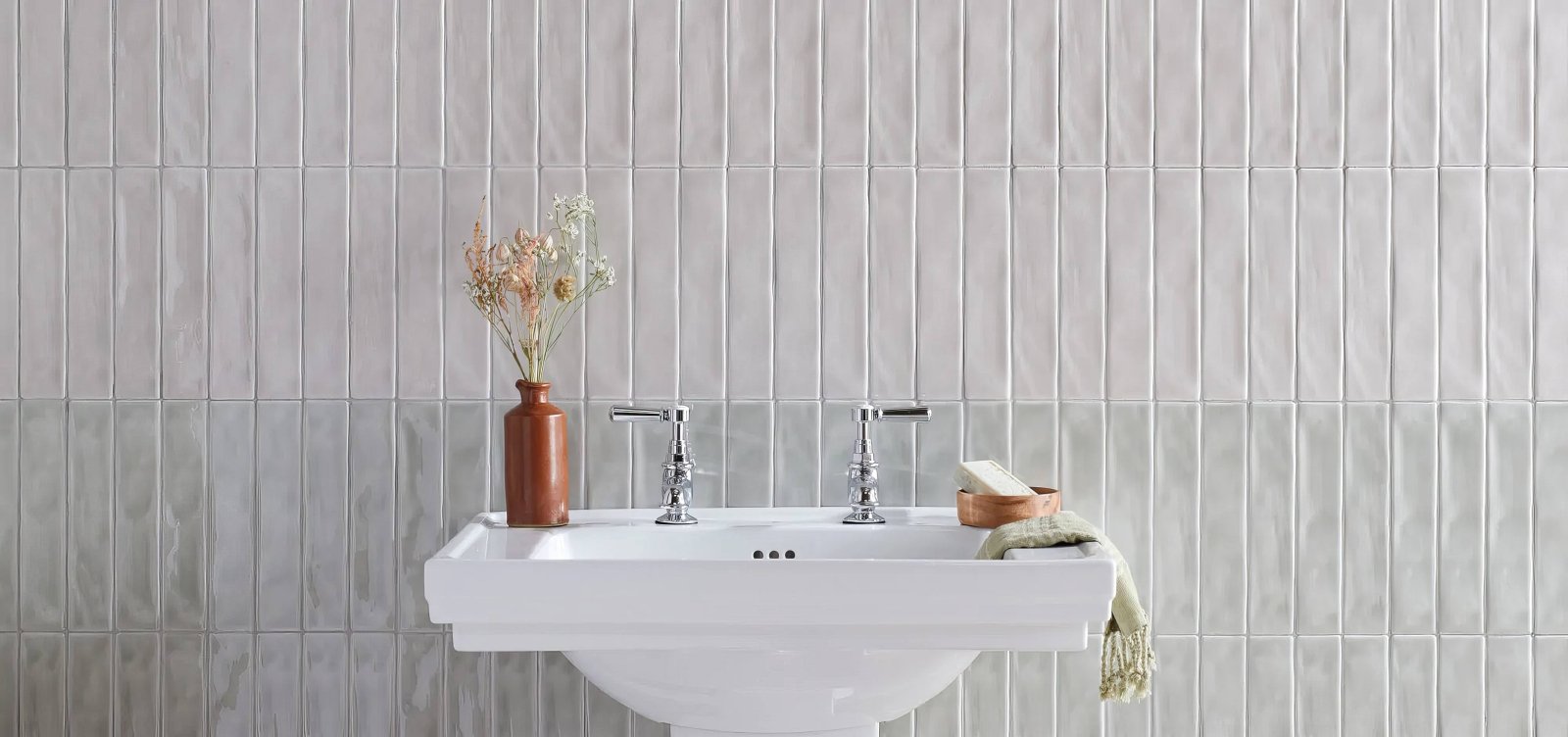 White ceramic pedestal sink with chrome faucets and a terracotta vase with dried flowers in onda tiles collection