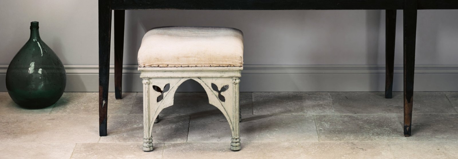 Antique-style stool with cream upholstered seat and distressed white gothic cutout legs in orient rustic floor tile setting