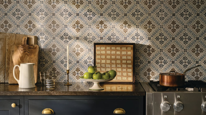 A white ceramic pedestal bowl filled with green apples on a dark countertop in patterned kitchen tiles collection