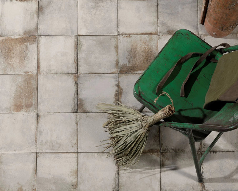 Rustic green metal wheelbarrow with weathered finish holding straw broom and brown straps in porcelain paving tiles collection