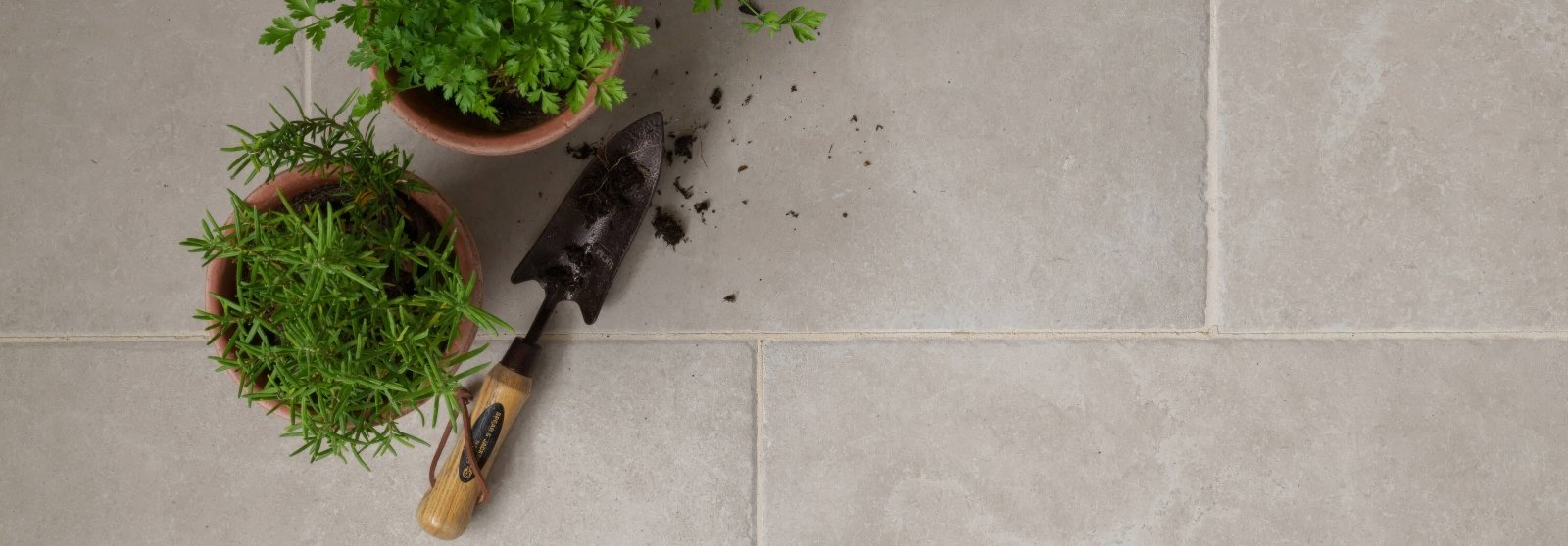 A well-used garden trowel with a wooden handle and metal blade beside two terracotta pots with green herbs in Quintili porcelain tiles.