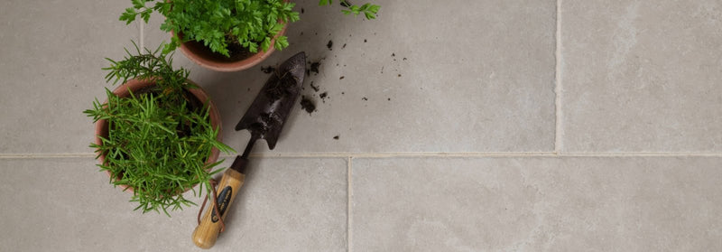 A well-used garden trowel with a wooden handle and metal blade beside two terracotta pots with green herbs in Quintili porcelain tiles.