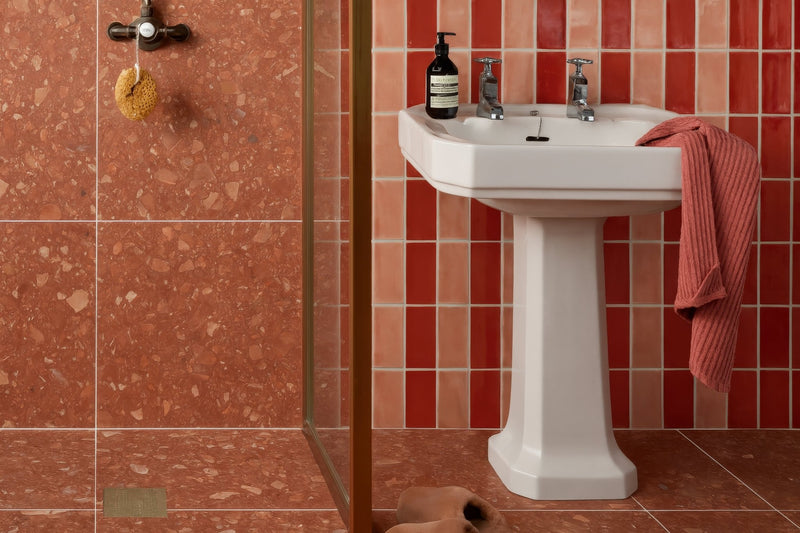 White ceramic pedestal sink with classic design and two chrome faucets in red bathroom tiles collection.