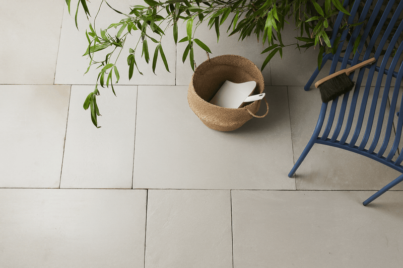 A woven natural fiber basket with two handles containing a white object resting on light gray sandstone floor tiles