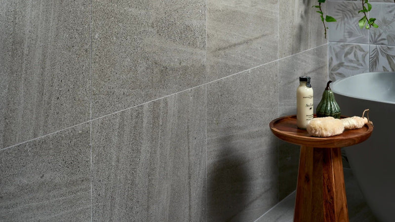 A round wooden side table with natural finish holding toiletries and decor in a sandstone wall tiles setting