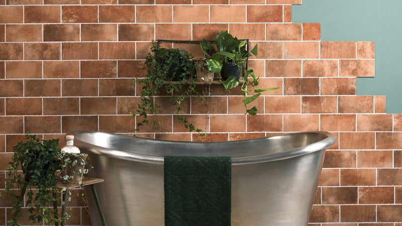 A sleek silver freestanding bathtub with polished metallic finish and a dark green towel in terracotta wall tiles setting
