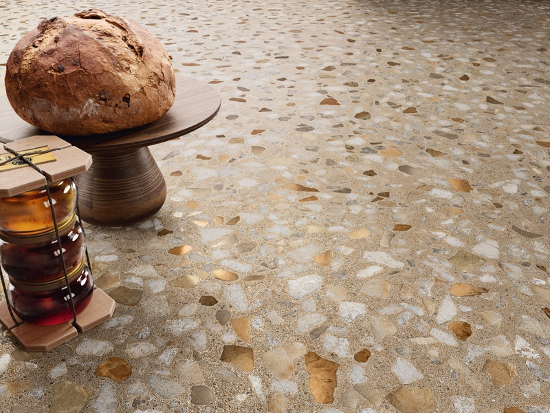 A rustic round artisan bread loaf with golden brown crust on a dark wooden pedestal in terrazzo effect tiles setting