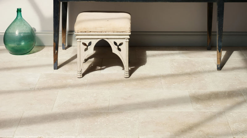 A cream-colored upholstered stool with ornate carved wooden legs and decorative arched base in travertine kitchen tiles collection.