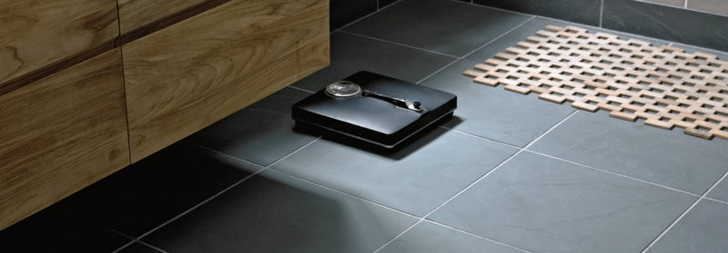 Black rectangular digital bathroom scale with circular display and silver-toned dial on urban slate tiles background