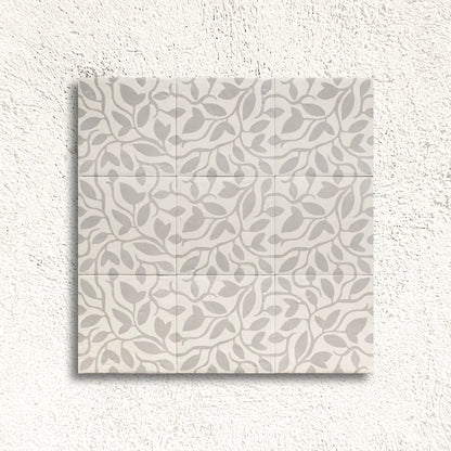 A square tile sample showing a repeating leaf pattern in light gray on a warm off-white background, mounted against a textured white wall.