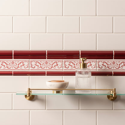 A glass bathroom shelf with gold hardware mounted on a white subway tile wall, featuring a red and white decorative tile border and a clear soap dispenser on the shelf.