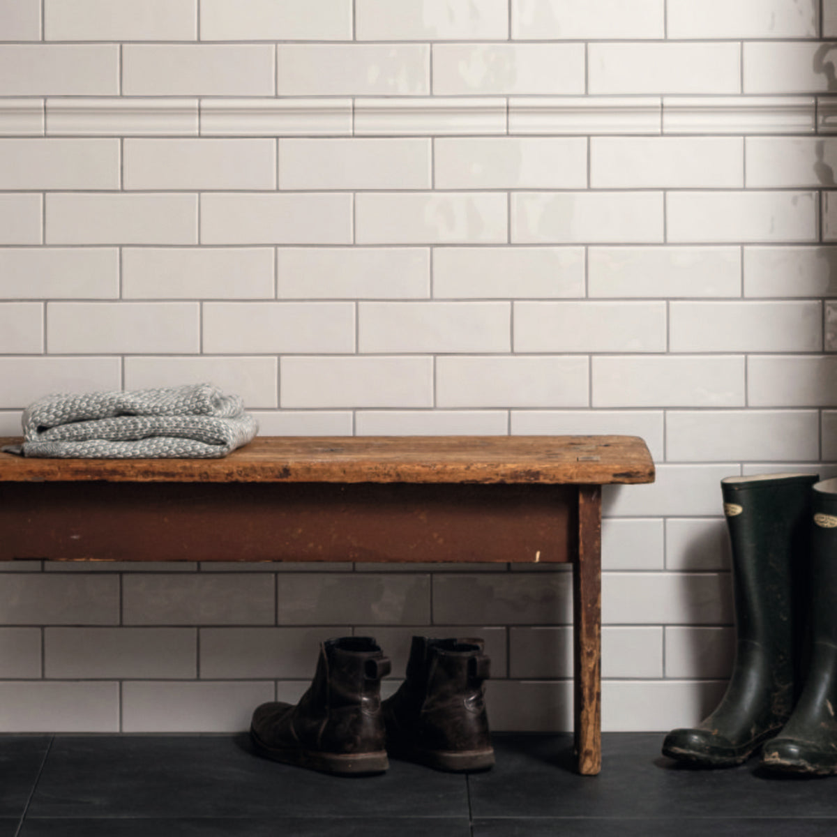 Wooden bench with folded clothes against a tiled wall, with boots and rain boots on the floor.