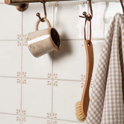 Beige mug, wooden brush, and checkered towel hanging on a rack against a tiled wall.