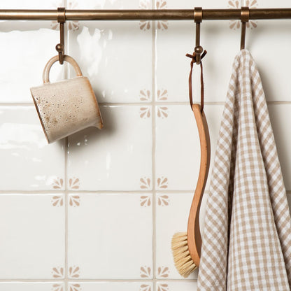 Ceramic mug, wooden spoon, and checkered towel hanging on a rack against a tiled wall.