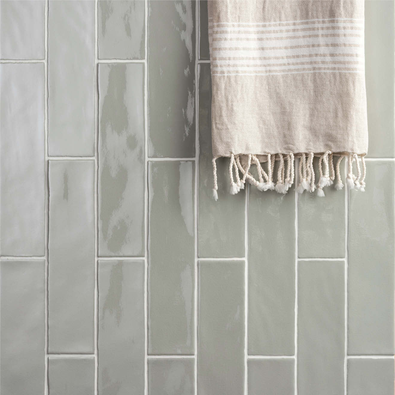 A soft beige towel with white horizontal stripes and fringed edges hanging against large brick tile tern gray backdrop