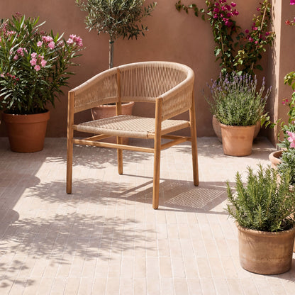 A light natural wood and woven rattan lounge chair placed on a tiled patio in a sunlit outdoor setting with potted plants and a terracotta wall.