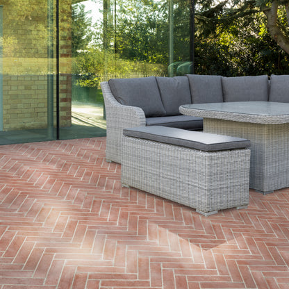 Gray wicker outdoor sectional furniture set with a matching ottoman and round table, arranged on a red brick patio in front of glass doors with greenery visible in the background.
