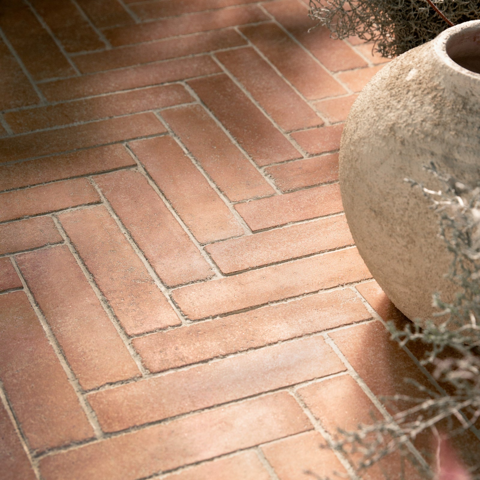 herringbone brick pavers arranged in a neat pattern, with a rustic beige stone planter partially visible on the right side.