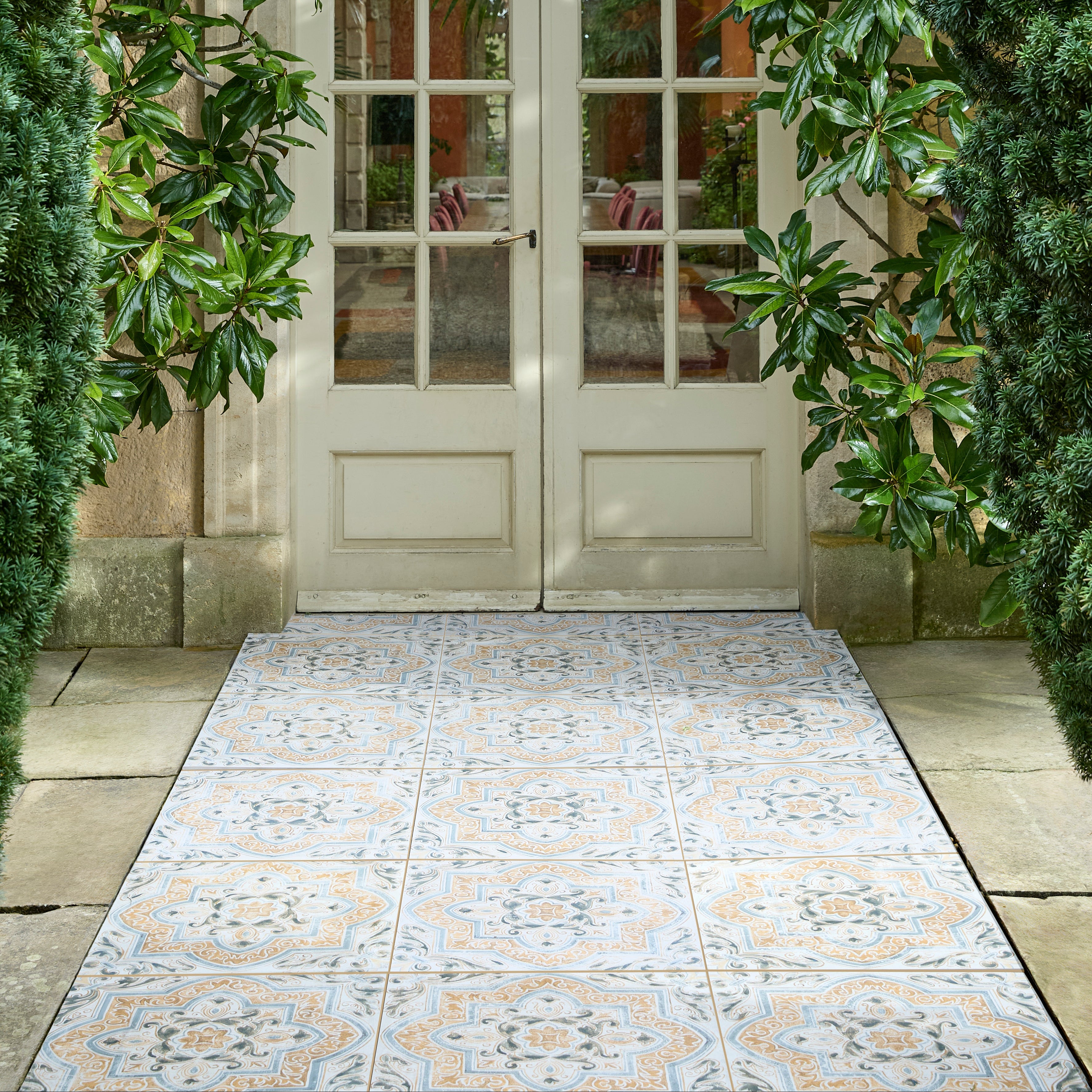 A pair of cream French doors with glass panes, framed by lush green foliage, with a decorative patterned tile runner leading up a stone path.