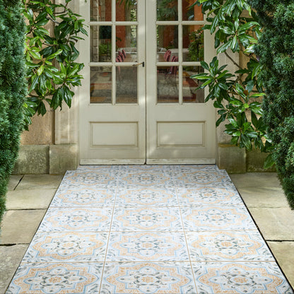 A pair of cream French doors with glass panes, framed by lush green foliage, with a decorative patterned tile runner leading up a stone path.