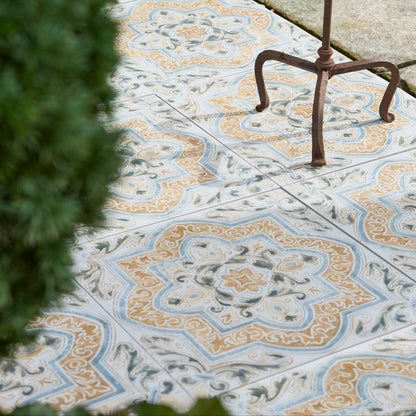 Decorative outdoor paving tiles with a blue, beige, and orange floral pattern, shown on a stone patio with a metal garden stand and green foliage.