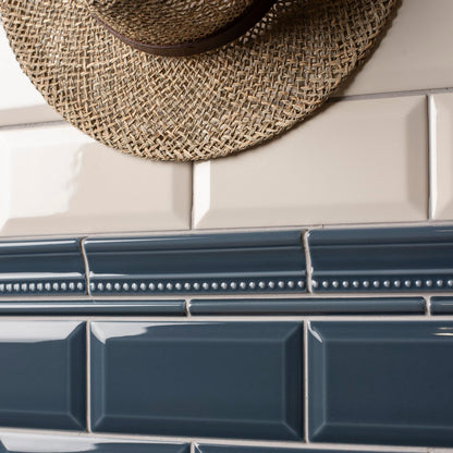 A close-up of glossy rectangular ceramic subway tiles in two colors: a dark slate blue lower section and a warm cream upper section, separated by a decorative white beaded tile band. A woven straw hat is partially visible at the top edge.