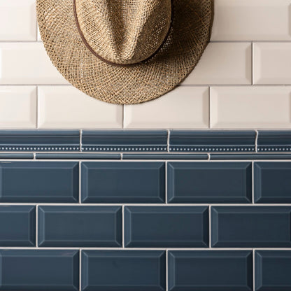 Two styles of subway tiles shown in a split layout: white glossy rectangular tiles on the top half with a woven straw hat hanging on the wall, and dark blue-gray glossy rectangular tiles on the bottom half.
