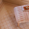 A soft pink and cream geometric-patterned upholstered ottoman with wooden legs in the adventuras ziggy corner terracotta porcelain design