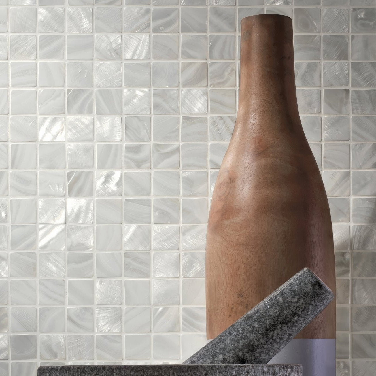 A tall slender wooden bottle with a smooth natural finish stands against white shimmering mosaic tiles in bianco 25mm square