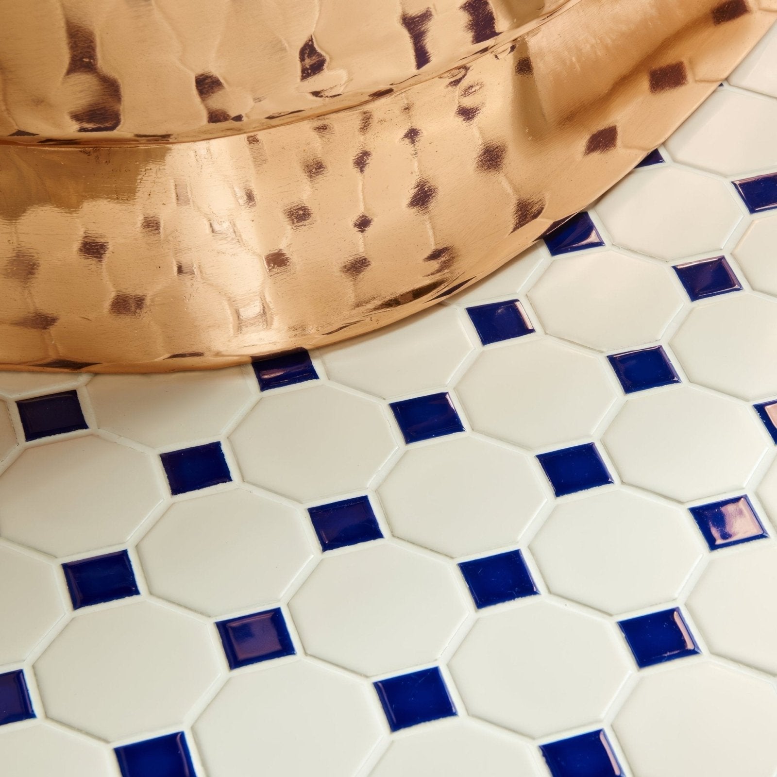 A hammered copper bowl with a warm reflective surface resting on a blue octagon and dot white hexagonal tile floor mosaic
