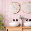 Two round mirrors with light wood frames and marble-like surfaces hang above white sinks on a blush smooth brick tile wall