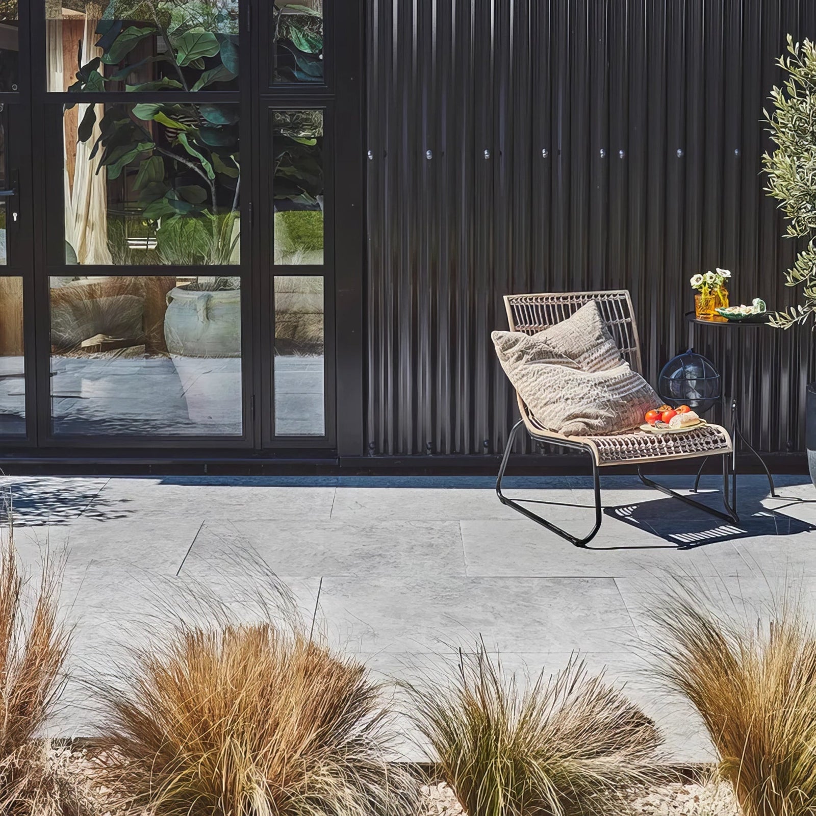 A beige woven rattan lounge chair with textured cushion and black metal frame outdoors on brit stone pennant porcelain paving