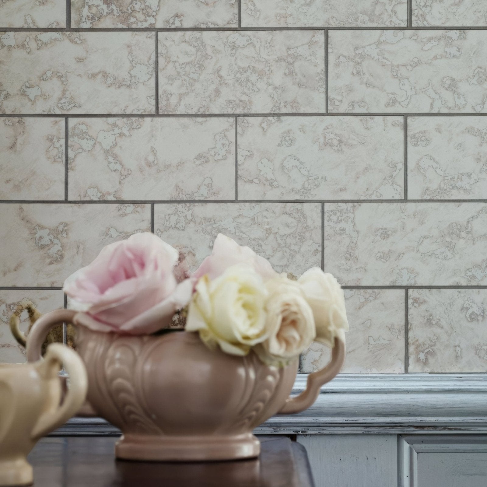 A beige ceramic vase with ornate embossed detailing and handles holding pink and cream roses in deco glass pewter tile