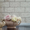 A beige ceramic vase with ornate embossed detailing and handles holding pink and cream roses in deco glass pewter tile