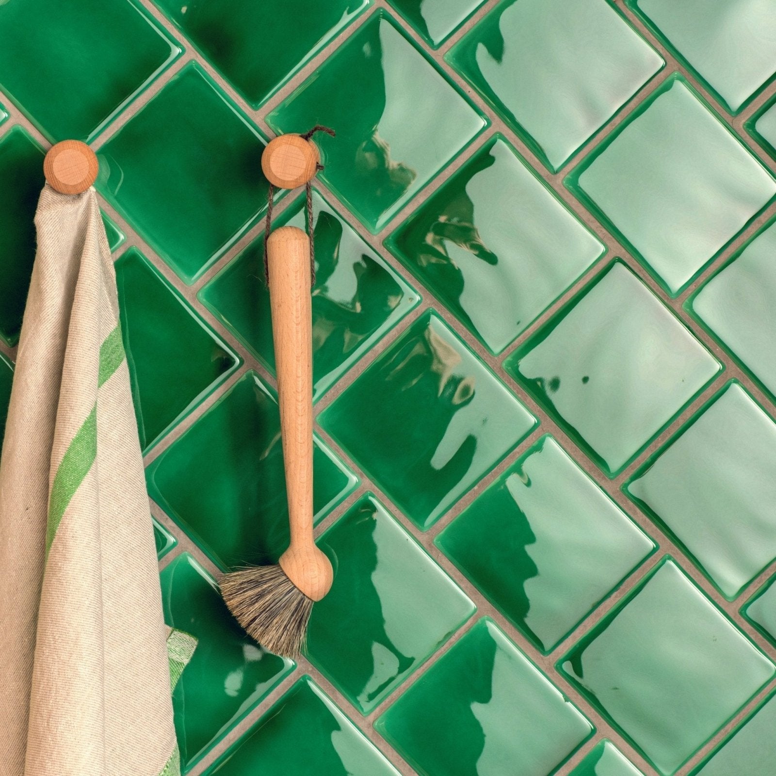 A wooden-handled scrub brush with natural bristles hangs on a wooden hook against emerald green field tile