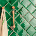 A wooden-handled scrub brush with natural bristles hangs on a wooden hook against emerald green field tile