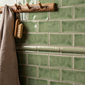 A rustic wooden towel rack with three natural-toned hooks mounted on glossy sage green ceramic half tile estuary wall