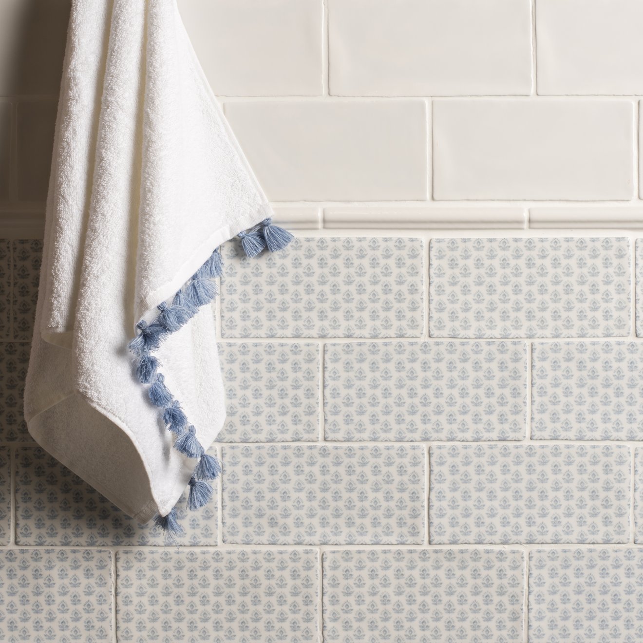 A soft white Fabrique Picot French Blue towel with delicate blue tassels hangs against a tiled wall