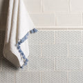 A soft white Fabrique Picot French Blue towel with delicate blue tassels hangs against a tiled wall