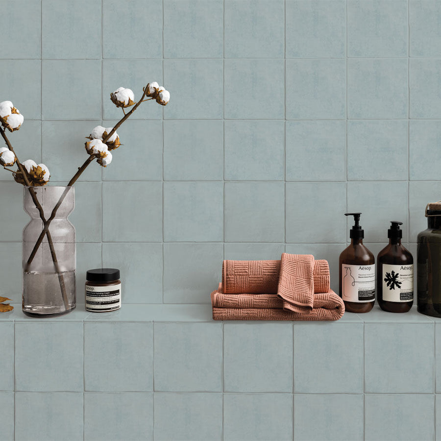 Bathroom shelf with towels, bottles, and a vase against a tiled wall.