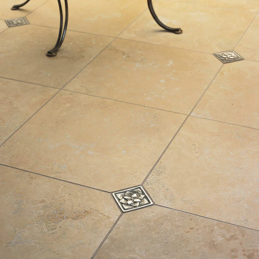 Square metallic inlay with raised floral design set into beige stone tiles from foundry lotus inset series