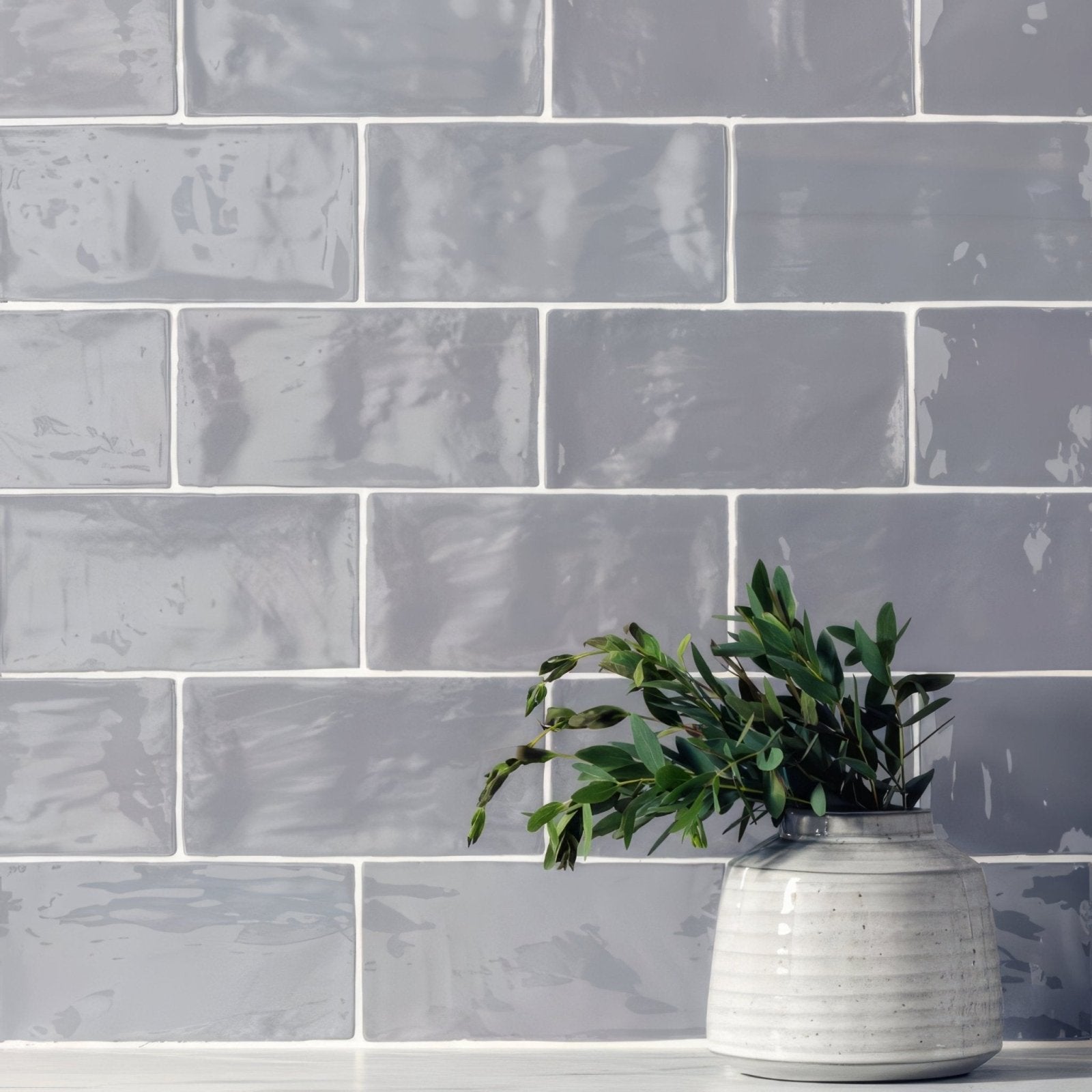 A white ceramic vase with horizontal ridges holding a green plant on a heather brick wall tile 20x10cm