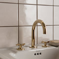 Gold-toned sleek curved faucet with cross-handle controls on white ceramic sink in helmingham crackle field tile setting