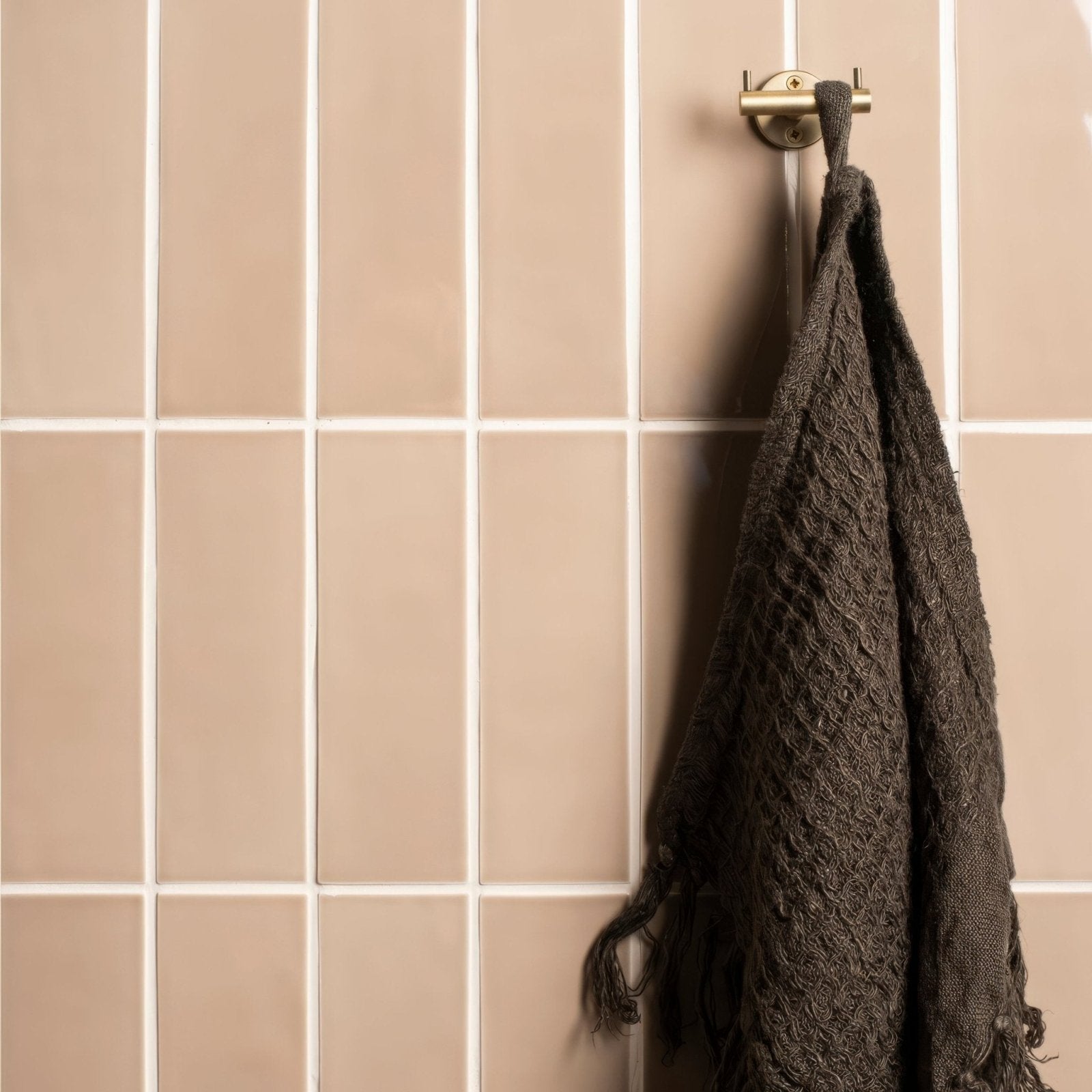 A dark brown textured knit towel with frayed edges hanging on a brass hook against beige brick tile ickworth walls