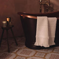 Cream-colored textured bath towel with tassels draped over a dark copper bathtub with jaipur akola patterned tile background