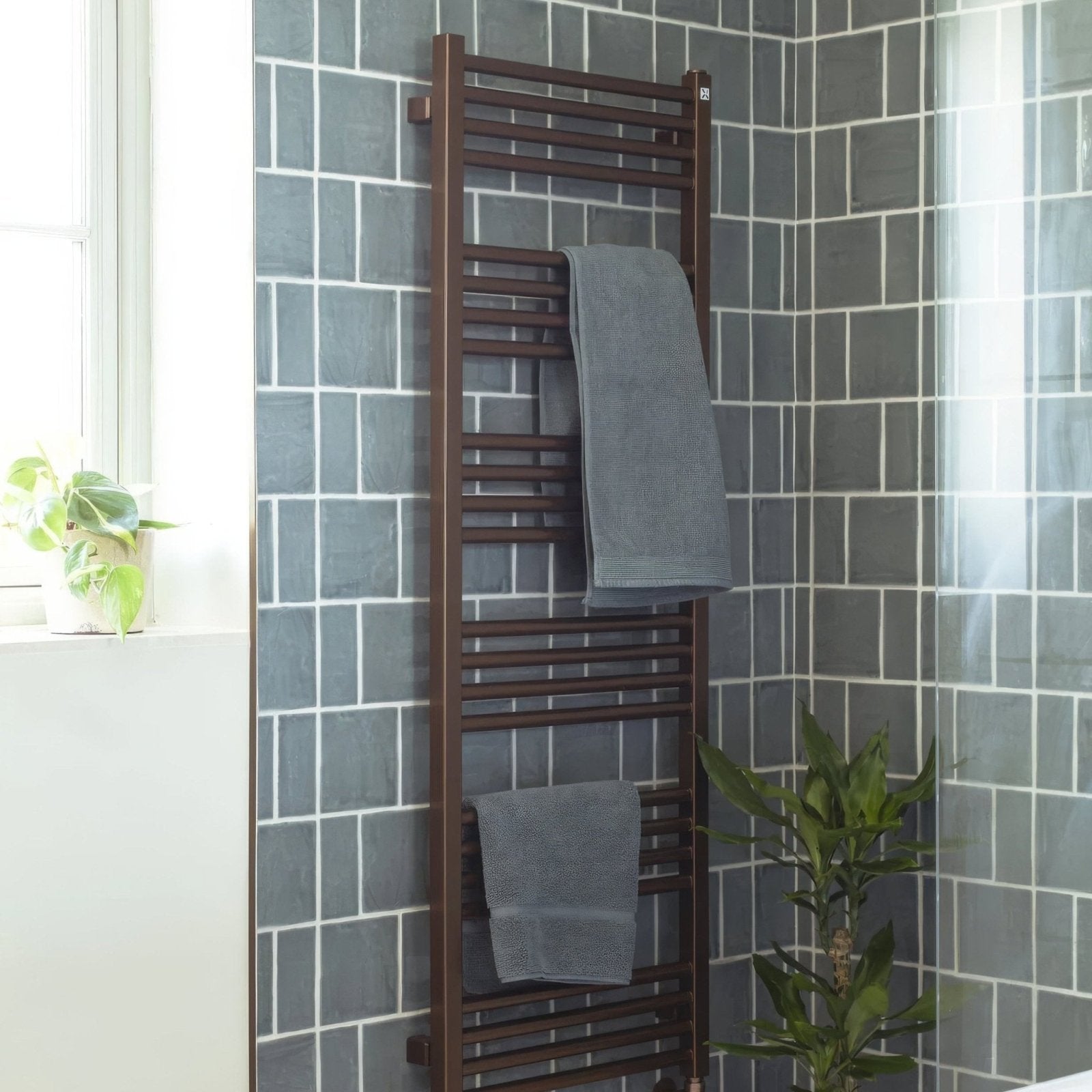 Brown heated towel rack with multiple horizontal bars holding two gray towels in front of jaipur dartmouth green tile