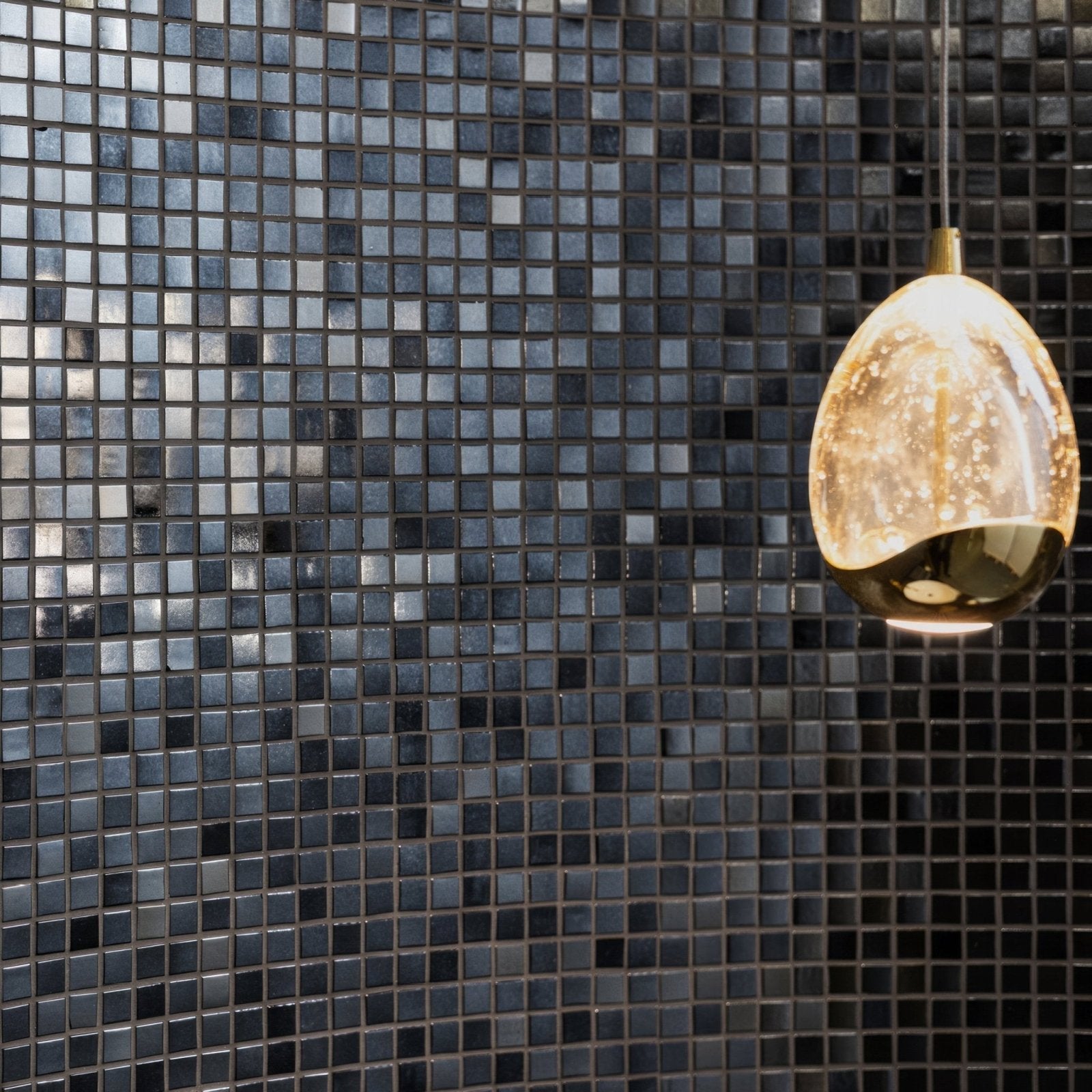 A sleek egg-shaped pendant light with warm glow and metallic gold accents hangs on a dark blue jungfrau iridescent glass mosaic wall